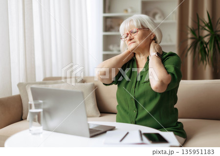 Senior woman massaging neck with eyes closed at home while using laptop 135951183