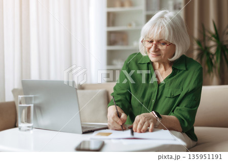 Senior woman writing notes at home while using laptop computer Senior woman writing notes at home while using laptop computer 135951191