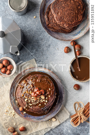 Chocolate pancakes topped with nuts and drizzled with chocolate syrup on a plate on gray textured background. Cozy breakfast and coffee, top view Chocolate pancakes topped with nuts and drizzled with chocolate syrup on a plate on gray textured background. Cozy breakfast and coffee, top view 135951892