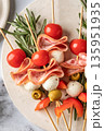 Snack skewers with mozzarella, ham, olives, peppers, tomatoes. Festive canapes or antipasti on white tray for new year table close up, top view 135951935