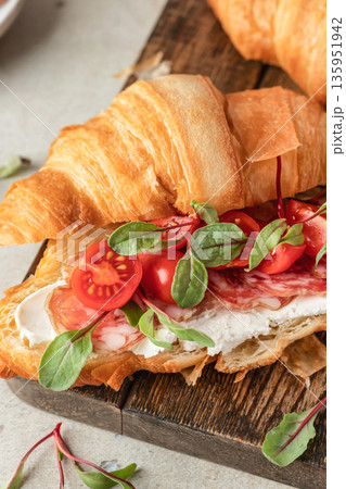 Croissant sandwich with salami, sausage, cherry tomatoes, soft cheese and chard micro greens on wooden board close up for savory healthy breakfast Croissant sandwich with salami, sausage, cherry tomatoes, soft cheese and chard micro greens on wooden board close up for savory healthy breakfast 135951942