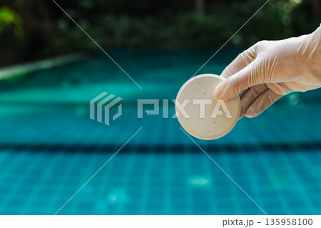 Gloved hand holding chlorine tablet over swimming pool water with blue tiles and soft blurred background, pool maintenance and sanitation concept  135958100