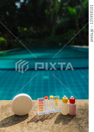 Pool water testing kit with chlorine tablet and reagent bottles on pool edge for swimming pool maintenance, chemical balance, and water quality control with copy space 135958101
