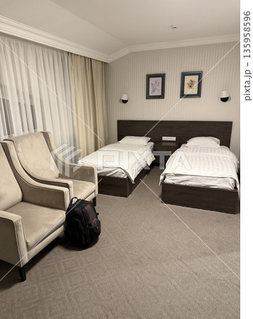 Hotel room with twin beds, armchair, and luggage, featuring neutral tones and minimal decor 135958596
