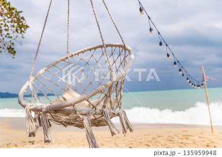 Hanging rope chair above sandy tropical beach with ocean waves and string lights, seaside relaxation scene with copy space for summer travel concept 135959948