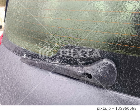 Close-up of car rear window wiper frozen in place by thick ice and frost, demonstrating harsh winter conditions affecting vehicle functionality. 135960668
