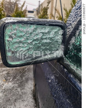 Close-up of icy green car side mirror encrusted in winter frost showing textured surface, frozen droplets and reduced clarity through glazed frozen glass. 135960677