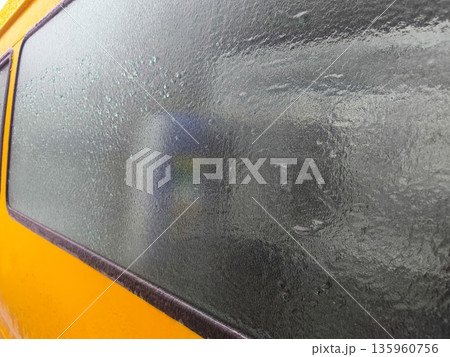 Frozen condensation completely covering yellow van window, creating textured icy surface and blurred view of interior objects through thick winter frost. 135960756