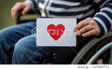 A disabled boy sits in a wheelchair and holds a Valentine's Day card. The concept is about Valentine's Day, inclusivity, love for others, and caring. A disabled boy sits in a wheelchair and holds a Valentine's Day card. The concept is about Valentine's Day, inclusivity, love for others, and caring. 135960772