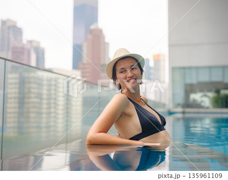 Young asian woman relaxing by pool at Kuala Lumpur hotel with view of surrounding skyscrapers, enjoying leisure time in vibrant urban setting. 135961109