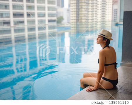 Young asian woman relaxing by pool at Kuala Lumpur hotel with view of surrounding skyscrapers, enjoying leisure time in vibrant urban setting. 135961110