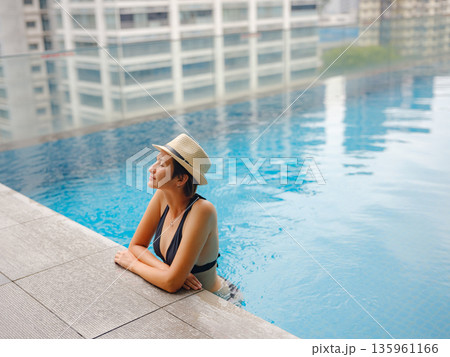 Young asian woman relaxing by pool at Kuala Lumpur hotel with view of surrounding skyscrapers, enjoying leisure time in vibrant urban setting. Young asian woman relaxing by pool at Kuala Lumpur hotel with view of surrounding skyscrapers, enjoying leisure time in vibrant urban setting. 135961166