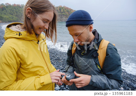 Couple enjoys Turkish coastline. walking in stone beach, watching sea waves, relaxing together, slow romantic moment near calm blue water under soft winter light. Couple enjoys Turkish coastline. walking in stone beach, watching sea waves, relaxing together, slow romantic moment near calm blue water under soft winter light. 135961183