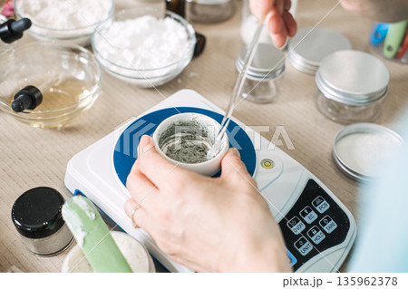Hands stir gray cosmetic paste in small bowl on digital scale surrounded by laboratory glassware and jars. Small-batch manufacturing, slow beauty movement, artisan production. 135962378