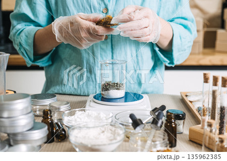 Person in protective gloves adds botanical ingredients to glass beaker on digital scale. Micro-manufacturing, artisan lab, small-scale cosmetics. 135962385