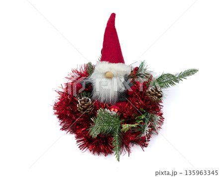 Cute Christmas gnome, featuring red hat and white beard, is nestled within tinsel wreath adorned with pine cones. This winter decoration provides cozy holiday atmosphere for any indoor space. Cute Christmas gnome, featuring red hat and white beard, is nestled within tinsel wreath adorned with pine cones. This winter decoration provides cozy holiday atmosphere for any indoor space. 135963345