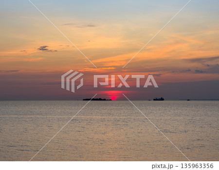 Stunning sunset casts vivid orange and red light across the calm water. Silhouetted land and ship line the distant horizon, creating peaceful maritime scene. Marine landscape background. 135963356