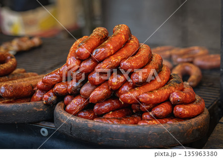Pile of sausages at food stand. Grilled sausage on tray, barbecue grill with fire heat and smoke, street food market stall. Concept of sausage street food cooking, catering service, food marketing 135963380