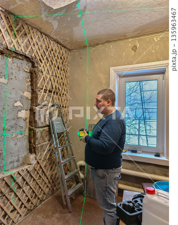 A man with a laser level projects green lines onto a wall with exposed wood siding and an unfinished ceiling while renovating an old room. A man is renovating a room in a house. 135963467