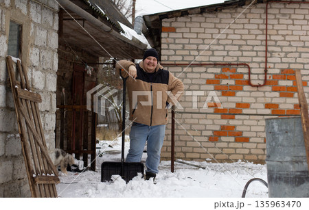 A man smiles while standing in a snowy yard, leaning on a snow shovel after clearing the paths. A happy man rests after clearing snow from his yard. 135963470