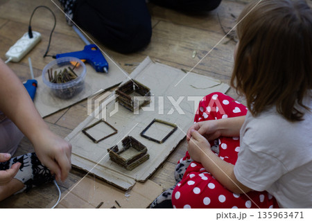 A mother and children sit on the floor crafting miniature houses out of wooden sticks using hot glue. The family is engaged in creative activities, learning, and enjoying the cozy atmosphere of home. 135963471