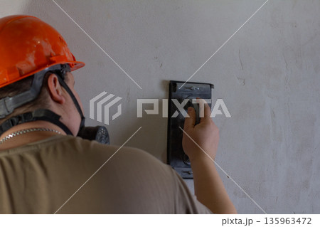 A construction worker wearing a safety helmet and a respirator mask carefully sands the rough surface of a wall with a manual grinder. A builder prepares a wall for painting during a house renovation. A construction worker wearing a safety helmet and a respirator mask carefully sands the rough surface of a wall with a manual grinder. A builder prepares a wall for painting during a house renovation. 135963472