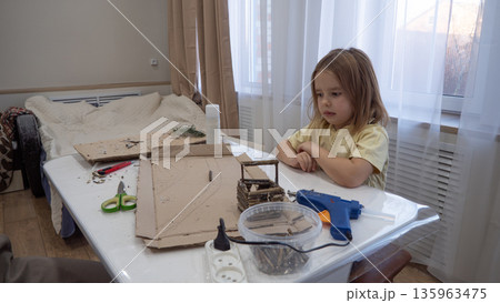 Child girl focused on DIY cardboard craft at table, using hot glue gun and scissors, doing hands-on learning and creative play at home. A child makes a craft from natural materials. 135963475