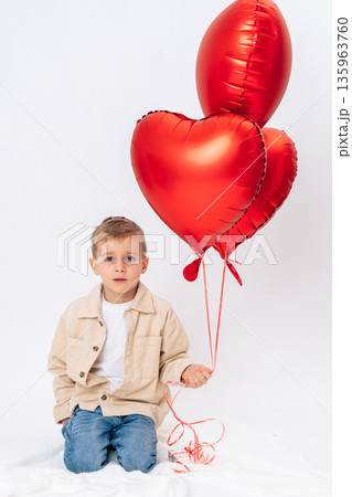 Little boy kneeling with red heart shaped balloons on white background. Valentines Day celebration, love symbol, childhood joy, studio portrait 135963760