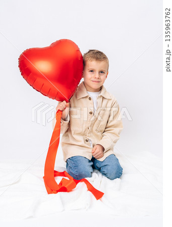 Little boy sitting and holding red heart balloon in front of face. Valentines Day, playful mood, love, childhood innocence Little boy sitting and holding red heart balloon in front of face. Valentines Day, playful mood, love, childhood innocence 135963762