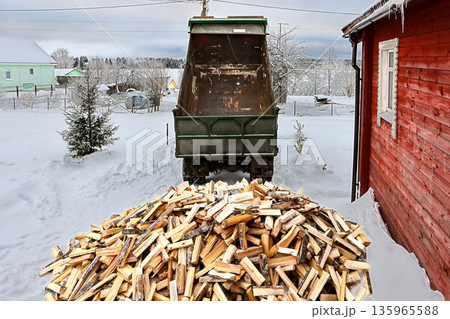 During heating season dump truck delivers firewood to houses with wood heating and unloads it onto snow in yard. 135965588