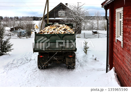 Dump truck prepares to unload split logs in front of rural house in snowy winter village. Dump truck prepares to unload split logs in front of rural house in snowy winter village. 135965590