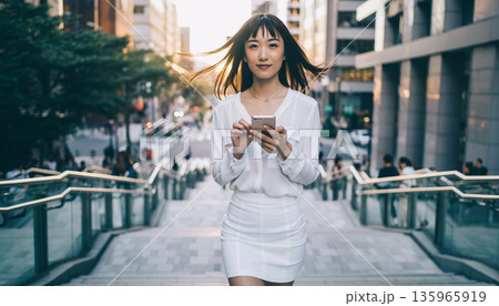 都会の街角でスマートフォンを手に空を見上げて微笑む20代の日本人女性 135965919