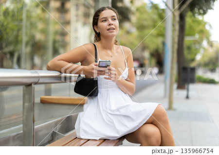 Positive young girl sitting at bus stop holding phone in hand 135966754