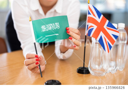 Female hands arranging flags of Saudi Arabia and United Kingdom on negotiating table 135966826