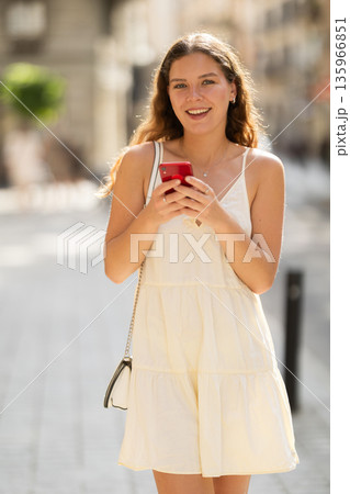 Happy young woman with mobile phone in her hands walking around city and taking pictures of sights 135966851