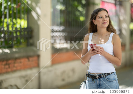 Happy woman looking to phone in hands and chatting in city Happy woman looking to phone in hands and chatting in city 135966872