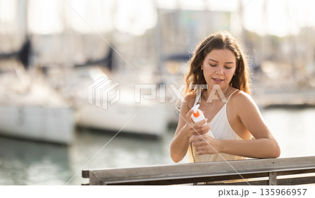 Young brunette girl applying sunscreen to her skin while walking in city 135966957