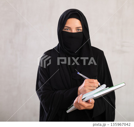 girl with face covered by black burka holds several notebooks and writes important note in diary girl with face covered by black burka holds several notebooks and writes important note in diary 135967042