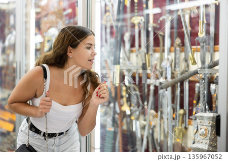 Enthusiastic young girl looking at handicraft swords in souvenir shop Enthusiastic young girl looking at handicraft swords in souvenir shop 135967052