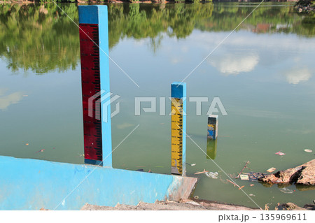 Water Level Markers indicating changes in a serene lake with reflection of green trees and sky under natural sunlight for environment monitoring Water Level Markers indicating changes in a serene lake with reflection of green trees and sky under natural sunlight for environment monitoring 135969611