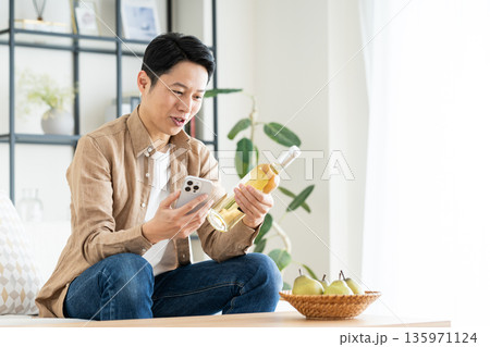 リビングでスマホとワインボトルを持つミドルの男性 135971124
