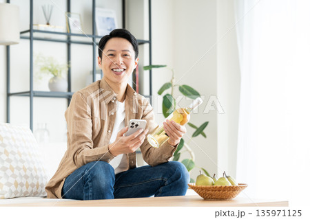 リビングでスマホとワインボトルを持つミドルの男性 135971125