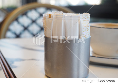 Sugar packets in a container next to a cup of coffee Sugar packets in a container next to a cup of coffee 135972990