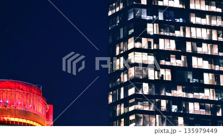 Big glowing windows in modern office building at night, in rows of windows light shines. Fragment of the glass facade of a modern corporate building at night. Modern glass office  in city. 135979449