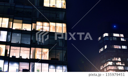 Big glowing windows in modern office building at night, in rows of windows light shines. Fragment of the glass facade of a modern corporate building at night. Modern glass office in city. Big glowing windows in modern office building at night, in rows of windows light shines. Fragment of the glass facade of a modern corporate building at night. Modern glass office in city. 135979451