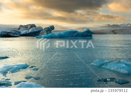Icebergs float in Iceland's glacier lagoon, reflecting the warm colors of sunset. Birds fly above, adding life to the tranquil scene. This location is a must-see for nature enthusiasts. 135979512