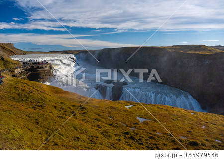 Gullfoss waterfall in Iceland showcases powerful cascades flowing into a canyon. Visitors admire the natural beauty from nearby viewpoints, especially during sunny weather. 135979536