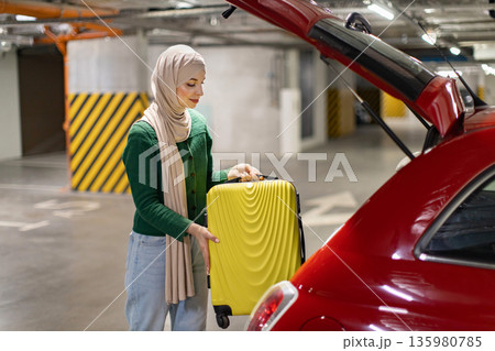 A woman wearing a hijab loads a yellow suitcase into the trunk of a red car in a parking garage A woman wearing a hijab loads a yellow suitcase into the trunk of a red car in a parking garage 135980785