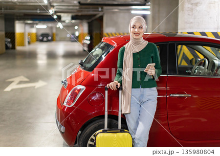 A woman wearing a hijab stands next to a red car in an underground parking garage, holding a phone and a suitcase A woman wearing a hijab stands next to a red car in an underground parking garage, holding a phone and a suitcase 135980804