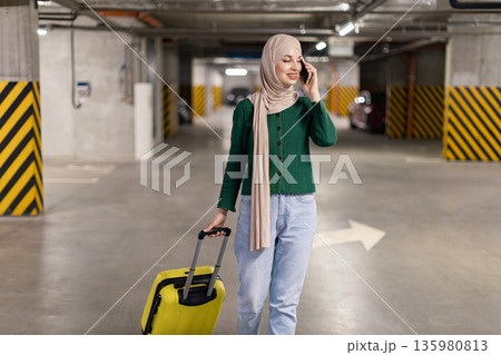 A woman wearing a hijab is talking on her phone while pulling a yellow suitcase in a parking garage A woman wearing a hijab is talking on her phone while pulling a yellow suitcase in a parking garage 135980813